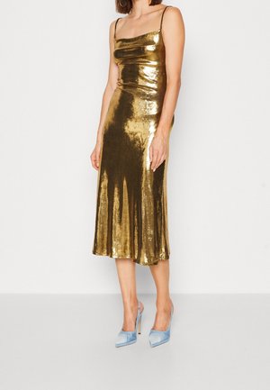 Nainen, joka pukeutuu kiiltävään kultaan vekattuun keskipitkään mekkoon, jossa on ohuet olkaimet, ja vaaleansiniset korkokengät, seisoo yksinkertaisen valkoisen taustan edessä.