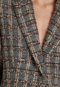 Blazer a cuadros gris y marrón que presenta una tela texturizada con un escote en V profundo. Patrón tejido distintivo con bordes deshilachados y un brillo sutil.