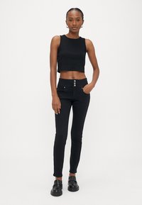 Mlada ženska stoji, oblečena v črno brezrokavnik crop top, črne hlače z visokim pasom in gumbi ter črne loaferje, na enostavni ozadju.