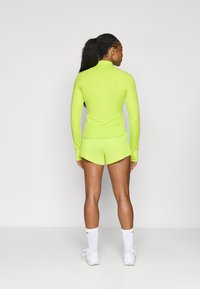 Conjunto deportivo en verde neón que incluye una camiseta de manga larga con un cuello alto y pantalones cortos elásticos con canalé. Combinado con calcetines tobilleros blancos y zapatillas.