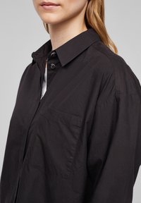 Svart skjorta med knappar och krage, en bröstficka och lös passform. Tillverkad av lätt tyg med en något strukturerad yta.