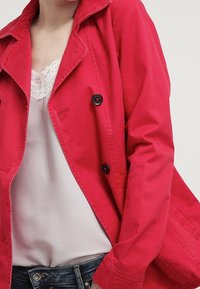 Gabardina rosa brillante hecha de tela ligera, con cuello, botones dobles y un ajuste relajado. Top de encaje blanco debajo.