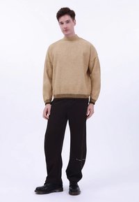 Maglione beige lavorato a maglia con collo alto e orlo a costine, abbinato a pantaloni scuri ampi e scarpe nere con lacci.