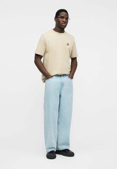 Άνδρας με μπεζ πουκάμισο με κοντά μανίκια, ανοιχτό μπλε τζιν παντελόνι φαρδύ, μαύρα παπούτσια και γυαλιά, στέκεται με τα χέρια στις τσέπες σε λευκό φόντο.