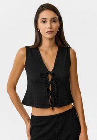 Zwarte, mouwloze, cropped top van een textuurstof, met een V-hals en een sluiting met een strik aan de voorkant. Het ontwerp heeft een losse, uitlopende zoom.