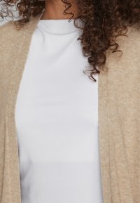 Beige stickad cardigan med öppen front och textilstruktur som hänger över en slät vit topp. Har subtila stygn och en avslappnad passform.