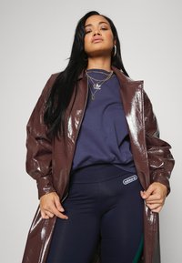 Blankett i glänsande brunt syntetmaterial över en marinblå sweatshirt, kombinerad med figurnära marinblå leggings med Adidas-logga. Guldhalsband i lager syns.