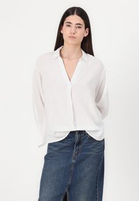 Camicia bianca di lino con collo, maniche lunghe e una vestibilità ampia, abbinata a una gonna di jeans blu scuro con spacco frontale.