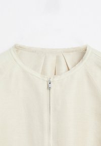 Beige textiel met een structuur, rond de hals, zichtbare stiknaden en een centrale zilveren rits.