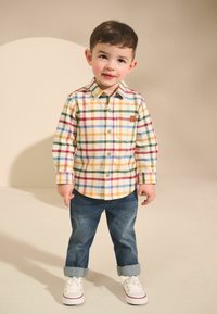 Chemise à boutons avec un motif écossais multicolore en jaune, vert, bleu et rouge ; associée à un jean en denim roulé et des baskets blanches.
