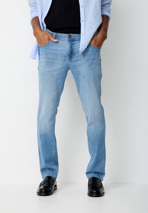 Mann steht mit den Händen in den Taschen, trägt hellblaue Jeans, schwarze Loafers, ein schwarzes Hemd und ein hellblaues, aufgeknöpftes Hemd.