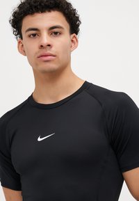 Cămașă sport neagră cu mânecă scurtă, din țesătură elastică, cu un logo alb Nike pe piept și mâneci raglan pentru mobilitate.