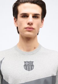 Νέος άνδρας με κοντά σκουρόχρωμα μαλλιά φορώντας ανοιχτό γκρι πουκάμισο με σκούρο λογότυπο FCB στο ύψος του στήθους, μπροστά σε λευκό φόντο.