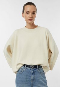 Cream sweater with a relaxed fit, ribbed neckline, and dropped shoulders, paired with blue denim jeans and a black belt featuring a silver buckle.