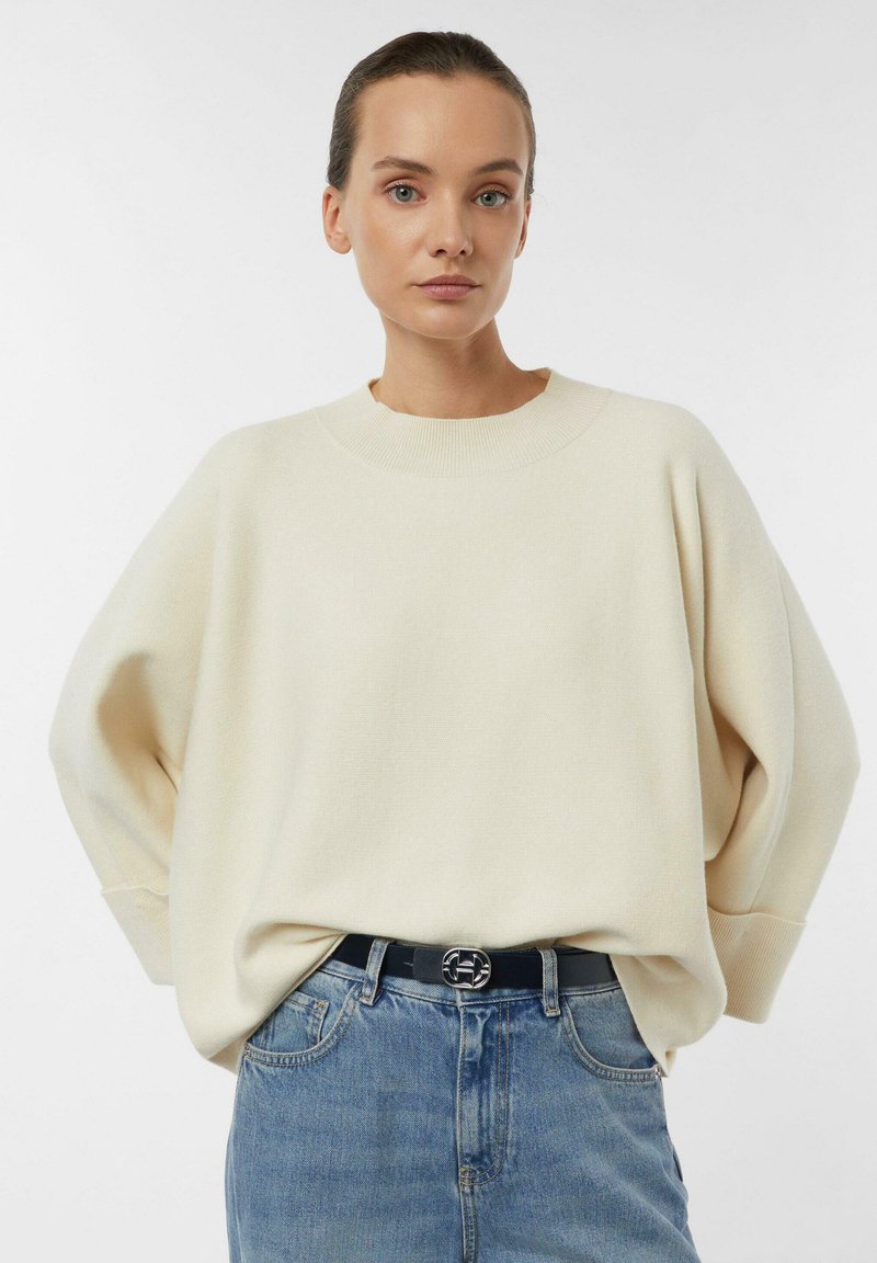 Cream sweater with a relaxed fit, ribbed neckline, and dropped shoulders, paired with blue denim jeans and a black belt featuring a silver buckle.