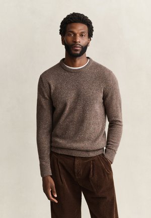 Homme avec barbe portant un pull marron moucheté et un pantalon en velours côtelé marron foncé, debout devant un fond clair uni.