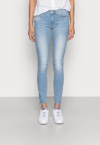 Mujer con jeans skinny de color azul claro con un leve desgastado y zapatillas blancas con el logo de Adidas en color lavanda, de pie sobre un fondo sencillo.
