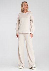 Jasno-beżowy zestaw dresowy składający się z luźnego swetra z opadającymi ramionami oraz spodni o szerokich nogawkach, wykonany z miękkiego, fakturowanego materiału.