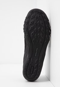 Semelle de chaussure Skechers noire avec motif de traction texturé et nom de la marque visible, positionnée verticalement contre un fond blanc en angle.