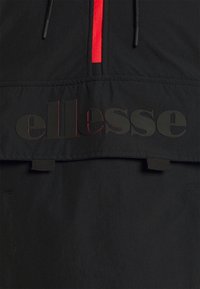 K-way nero con accenti rossi nella zip. Presenta un grande logo in rilievo nero sul davanti. Realizzato in tessuto leggero e testurizzato.
