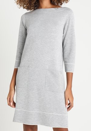 Lichtgrijze, knie-lange gebreide jurk met driekwart mouwen en twee opgestikte zakken aan de voorkant, gedragen door een persoon met krullend haar.