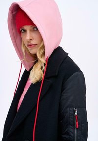 Blond žena nosí červenou čepici, růžovou mikinu s kapucí, černý kabát s prošívanými rukávy a dívá se přímo před sebe na bílém pozadí.