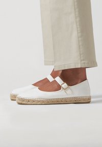 Des espadrilles blanches avec un bout rond, une sangle à boucle et une semelle en jute tissée. Le matériau supérieur est lisse, complété par une jambe de pantalon beige.