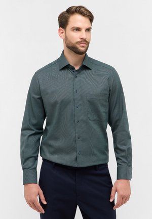 Camicia a maniche lunghe a quadri verde scuro con colletto a serratura, taschino frontale e bottoni blu, indossata con pantaloni scuri.