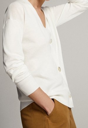 Osoba ubrana w biały kardigan na guziki z długimi rękawami i brązowe spodnie, z jedną ręką w kieszeni, a drugą uniesioną przy głowie.