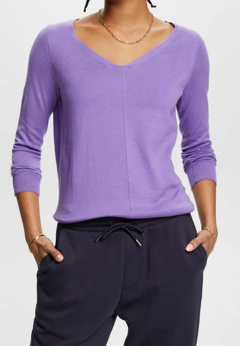 Personne portant un pull lavande à col en V et manches longues ainsi qu'un pantalon foncé à cordon, debout les mains dans les poches devant un fond uni.
