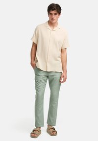 Camisa de botones de manga corta, color beige claro, con un patrón texturizado, combinada con pantalones ajustados verdes claros y sandalias de verano marrones.