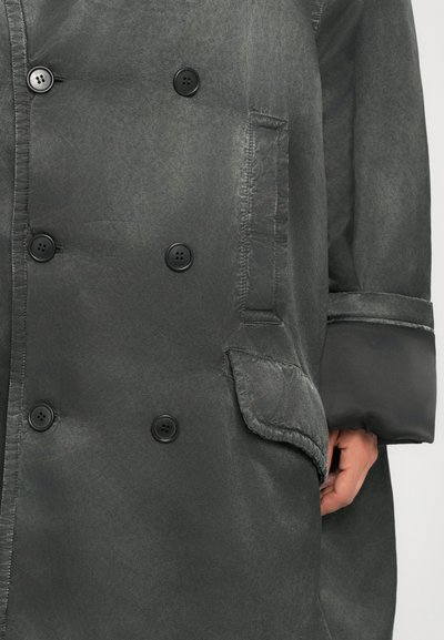 Σκούρο γκρι διπλόχρωμο παλτό με τέσσερα μαύρα κουμπιά, πλαϊνή τσέπη με καπάκι και μανίκι με γαζιά, που δείχνει εν μέρει ένα χέρι.