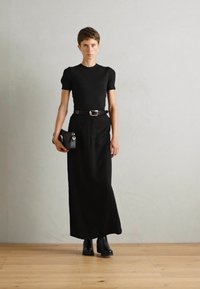 Crna, prilagođena kratka majica s kratkim rukavima u kombinaciji s dugom crnom suknjom, naglašena remenom. Model drži malu crnu torbu i nosi crne čizme.