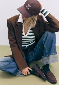 Brun blazer over en stribet hvid og grøn top, blå denim jeans med opbukkede kanter, og mørkebrune loafers. Kasketten står der "STOL PÅ PROCESSEN."