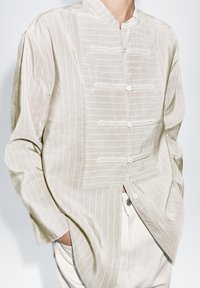 Jasnobeżowa koszula z kołnierzykiem typu stójka, teksturowanymi paskami i plisą na guziki. Luźny krój z długimi rękawami, wykonana z miękkiego, lekkiego materiału.
