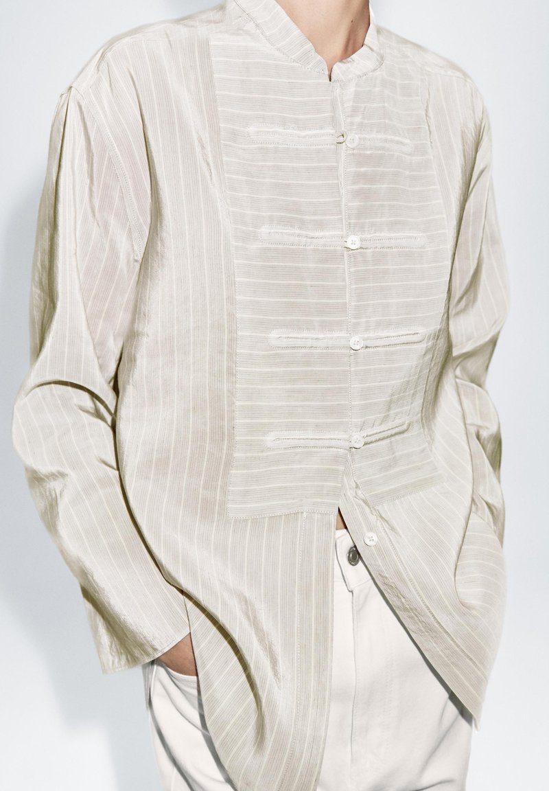 Jasnobeżowa koszula z kołnierzykiem typu stójka, teksturowanymi paskami i plisą na guziki. Luźny krój z długimi rękawami, wykonana z miękkiego, lekkiego materiału.