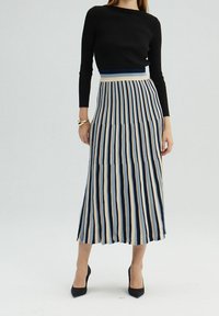Haut noir à manches longues à côtes associé à une jupe maxi plissée avec des rayures verticales en bleu marine, beige et bleu clair. Des escarpins noirs complètent la tenue.
