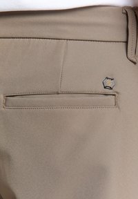 Lichtbruine broek met een gladde textuur, een achterzak en een hexagonaal logo-accent. Geometrisch ontwerp op de zak.
