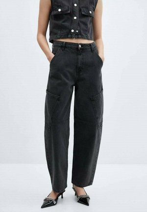 Vrouw draagt zwarte jeans met hoge taille en wijde pijpen, een mouwloos zwart kort gilet met knopen en zwarte puntige hakken met enkelbandjes.