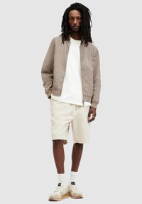 Lätt taupe bomberjacka över en vit skjorta, kombinerad med beiga shorts. Har kraftiga beiga sneakers och vita strumpor.