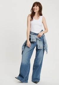 Biała bluzka na ramiączkach zestawiona z niebieskimi dżinsami z wysokim stanem i szerokimi nogawkami. Dżinsowa kurtka jest przewiązana w talii. Dżinsy mają przetarty wygląd.