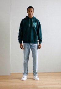 Felpa verde scuro con tasca frontale e stampa grafica; jeans azzurri chiari; sneakers bianche; colori solidi e design casual.