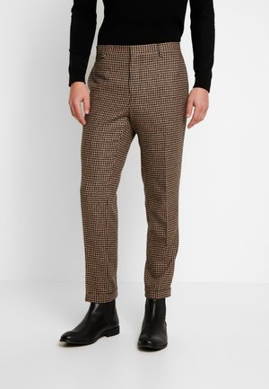 Mężczyzna w brązowo-czarnych, obcisłych spodniach w jodełkę, czarnych botkach za kostkę i czarnym, długim rękawem bluzce.