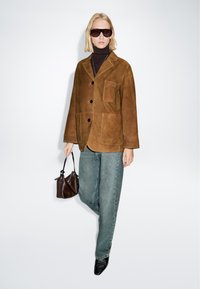 Giacca in suede marrone con grande tasca frontale, bottoni neri, indossata sopra un dolcevita scuro. Abbinata a jeans azzurri chiari e una piccola borsa marrone.