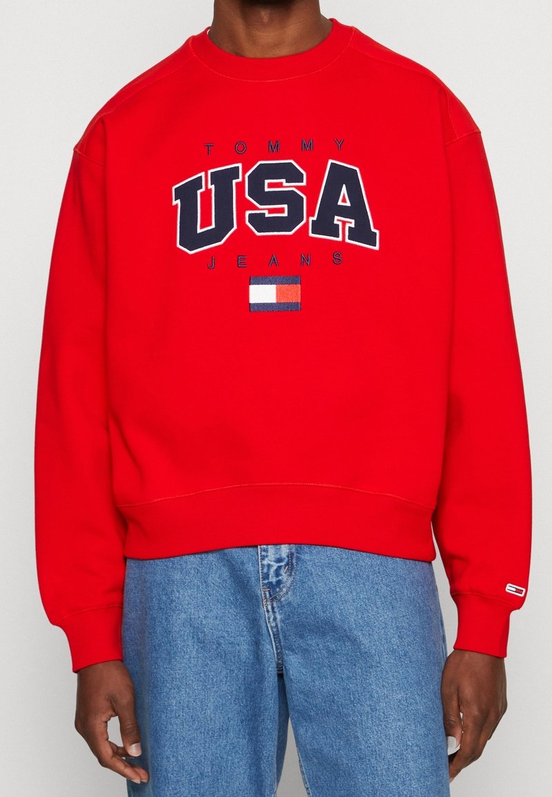 Sweat-shirt rouge avec l'inscription "TOMMY USA JEANS" en bleu marine, comprenant un petit graphique de drapeau. Matière en coton doux, design classique à col rond.