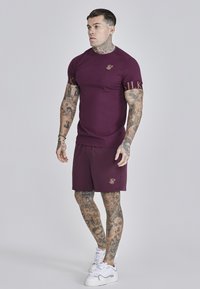 Maroonowa koszulka sportowa z złotym logo na rękawie, dopasowana do maroonowych szortów, oba elementy wykonane z gładkiego materiału, noszone z białymi sneakersami.