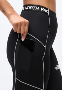 Χέρι τοποθετεί το smartphone στην πλαϊνή τσέπη μαύρου κολαν The North Face με λευκή ραφή και ζώνη μέσης με λογότυπο.