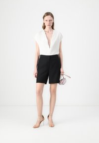Hvit, kortermet topp med glidelås, kombinert med skreddersydde svarte shorts. Modellen holder en liten, lys rosa håndveske og har på seg nude høyhælte sandaler.