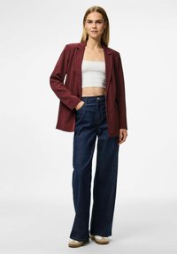 Woman wearing a white crop top, burgundy pinstripe blazer, dark wide-leg jeans, and beige sneakers, standing with one hand in pocket.