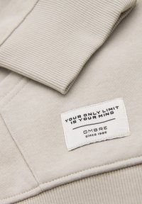 Poignet en tissu côtelé beige et étiquette blanche cousue sur le vêtement portant l'inscription "Votre seule limite est votre esprit" et "OMBRE depuis 1985".